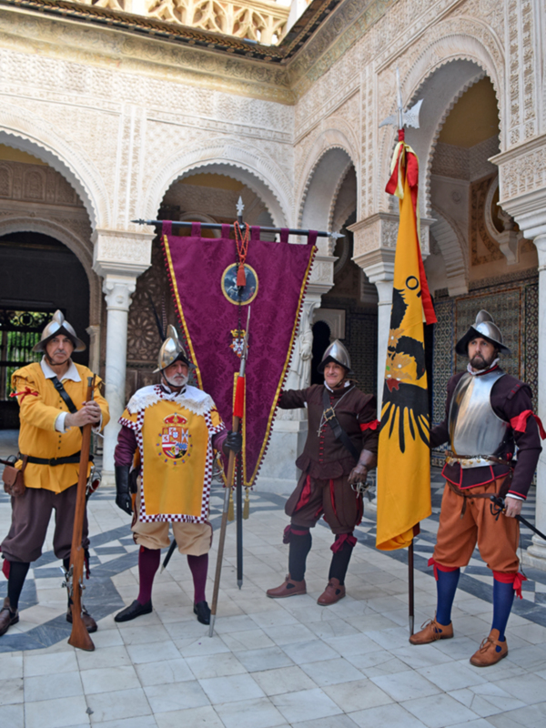 Guerreros medievales en un palacio histórico, portando armas y banderas, posando en un patio con arcos y decoración arquitectónica.