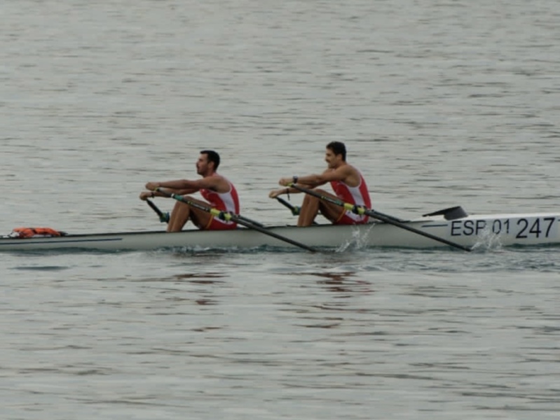 Dos hombres remando en una canoa de remo en el agua. La canoa tiene la marca 'ESF 01 247' en la parte delantera.