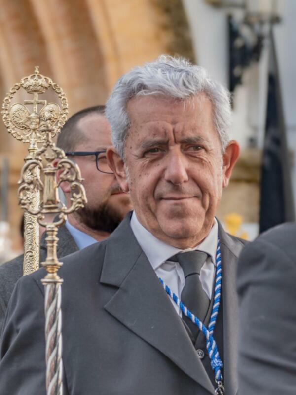 Un hombre con cabello gris y barba, vestido formalmente, sostiene una cruz dorada. Detrás de él, otros hombres también en traje. El fondo muestra una estructura arquitectónica con detalles de madera y un cielo claro.