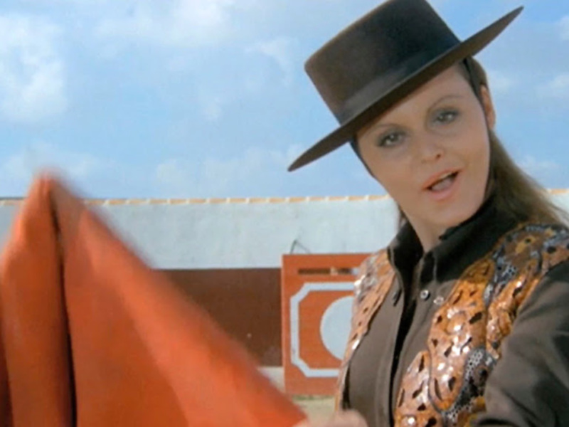 Una mujer vestida con un traje de vaquero y un sombrero, sosteniendo una bandera naranja en un entorno rural con un cielo azul y nubes blancas. La imagen sugiere un ambiente de acción o aventura, posiblemente en un entorno rural o western.