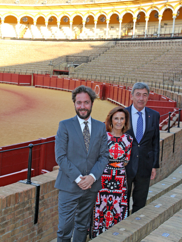 Dos personas en trajes posan frente a una arena de toros vacía. La imagen muestra detalles arquitectónicos y la textura del suelo de arena, enfatizando el contexto histórico y cultural de la imagen.