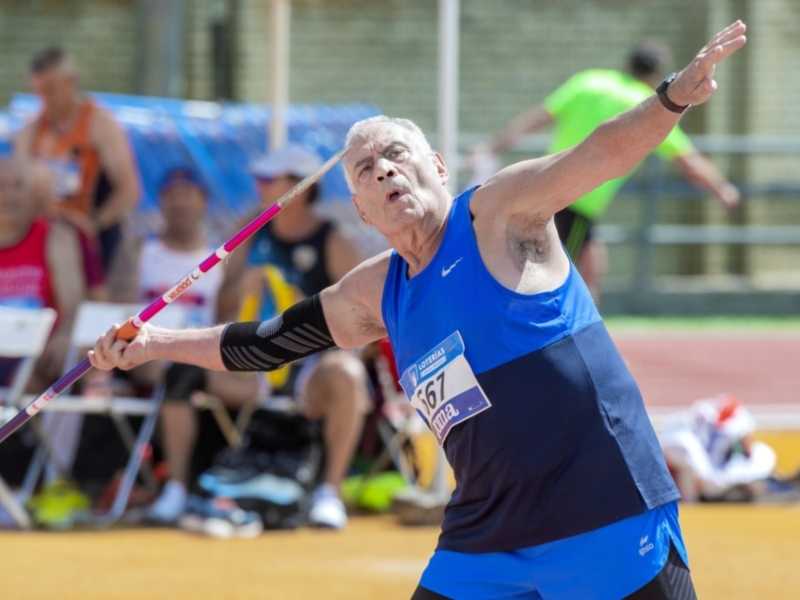 Un atleta en acción lanzando un javelín, con otros competidores y espectadores en el fondo.