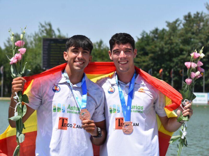 Dos atletas celebran su victoria en un evento deportivo, sonrientes y con medallas de oro y plata, uno de ellos sostiene un ramo de flores y una bandera española.