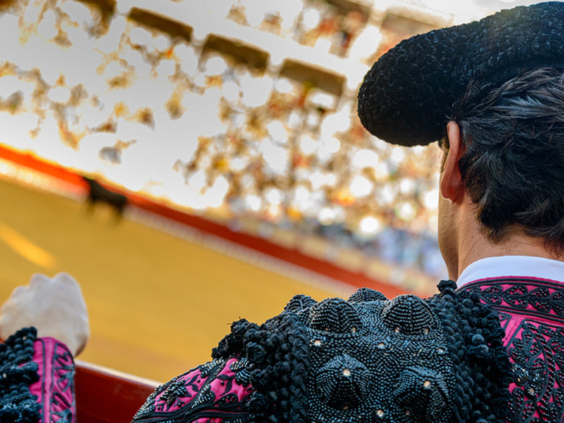 Un torero con una capa de color negro y rojo, observando la plaza de toros desde el lado derecho.