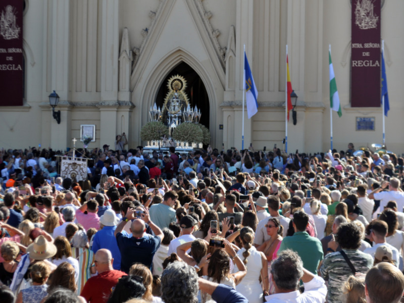 Una multitud de personas congregadas frente a una iglesia grande y alta, con un altar adornado en el centro. Banderas ondean en el aire, y la imagen transmite un ambiente festivo o religioso.
