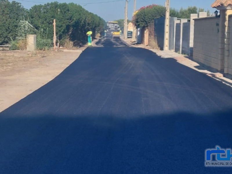 Carretera recién asfaltada en un pueblo, con árboles y viviendas a ambos lados.