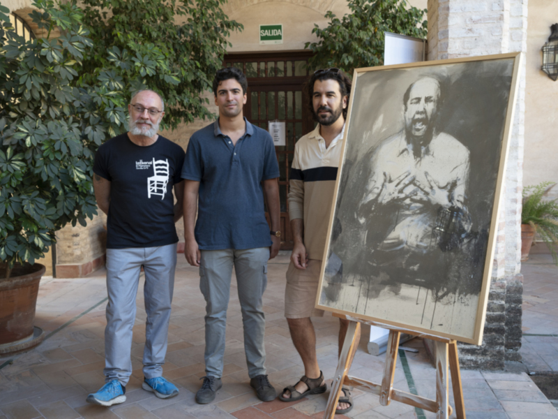 Tres hombres posan junto a una imagen de un hombre antiguo, en un entorno con plantas y una lámpara.