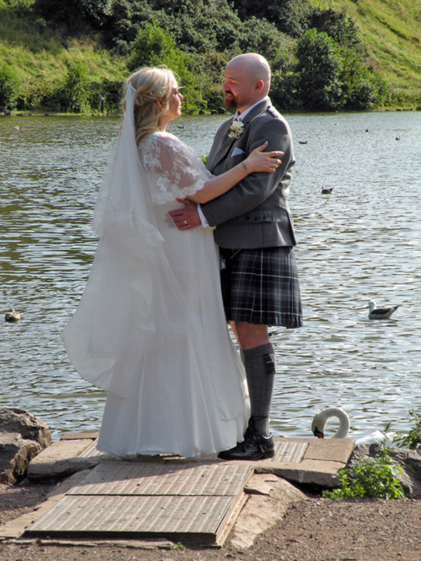Boda en Escocia con vestido blanco, pantalón escocés y novio con corbata. Lago con patos y vegetación en el fondo.