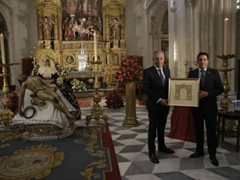 Un hombre en traje negro sostiene un lienzo con un diseño artístico en una iglesia, frente a un altar con una imagen religiosa y flores. La decoración del suelo es geométrica en azul y blanco, con un escudo en el suelo.