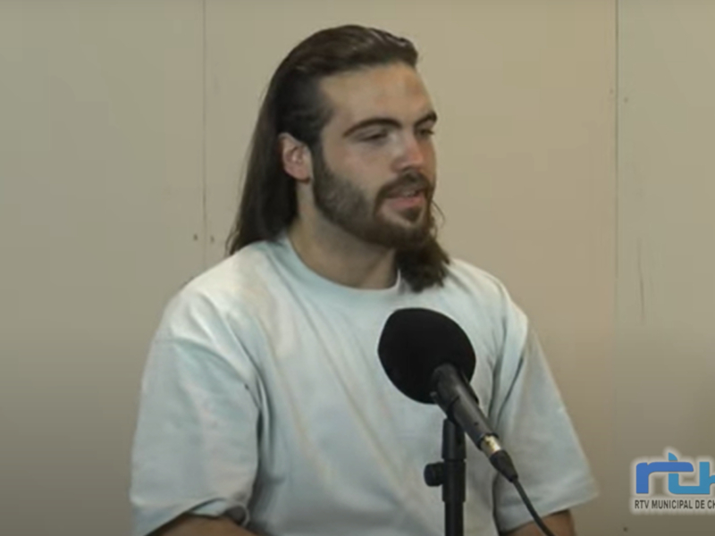 El hombre tiene una larga barba y cabello largo, está hablando en un micrófono.