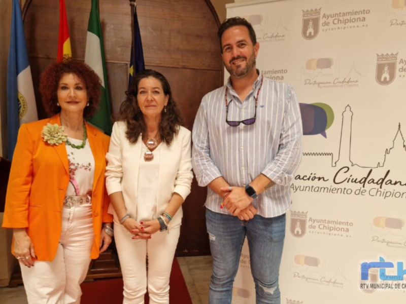 Se muestra a tres personas posando frente a una bandera de España y un cartel del Ayuntamiento de Chipiona. La persona en el centro lleva una camisa blanca y pantalones blancos, mientras que las otras dos están vestidas con prendas de color más oscuro. La escena parece ser una presentación o evento oficial, posiblemente relacionado con el municipio de Chipiona.