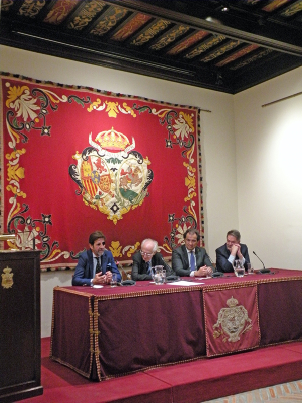 Conferencia de prensa en un salón con decoración histórica y escudo de armas en la pared.