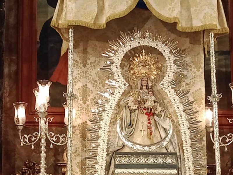 Elegante imagen religiosa de una Virgen María vestida con una túnica blanca y un manto dorado, rodeada de una corona de luz y adornada con flores y joyas. Está colocada en un altar con velas encendidas a su lado.