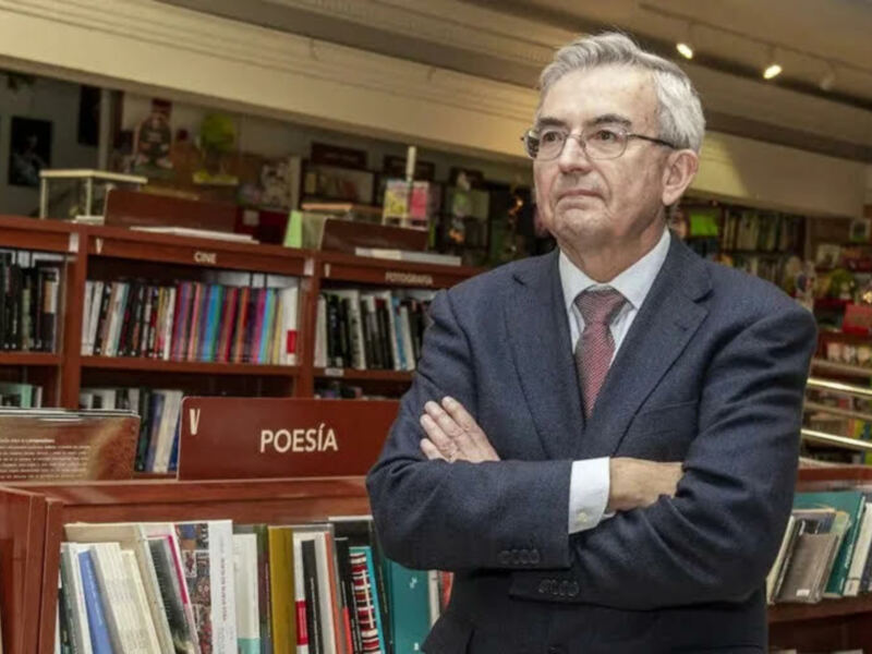 Un hombre en traje y gafas, con los brazos cruzados, frente a una estantería llena de libros en una librería.