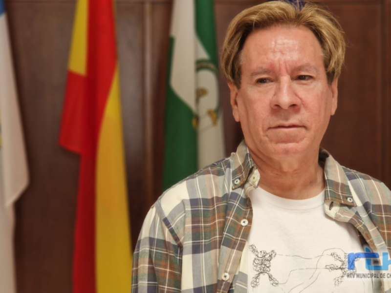 A person standing in front of flags, wearing a plaid shirt over a white t-shirt.