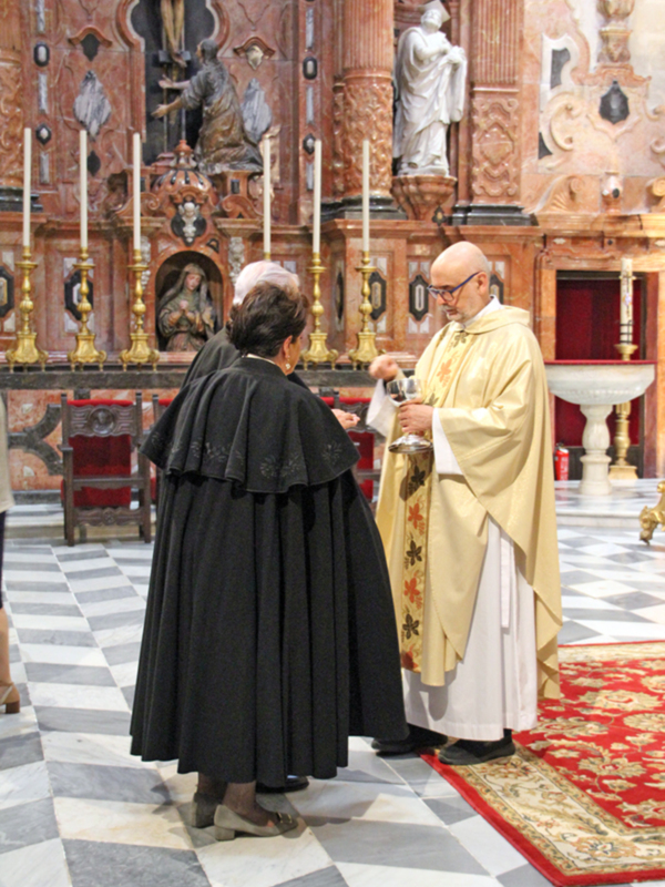 Una ceremonia religiosa en un templo con una mujer en traje negro y un hombre en vestido de sacerdote, ambos en actitud reverente. El fondo muestra un altar con estatuas y una decoración de marmol rojo y blanco.