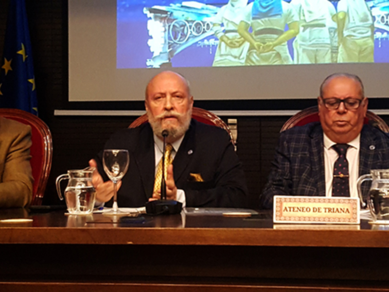 Tres hombres sentados en una mesa de conferencia, con banderas y un cartel que dice "Ateneo de Triana" en el fondo.