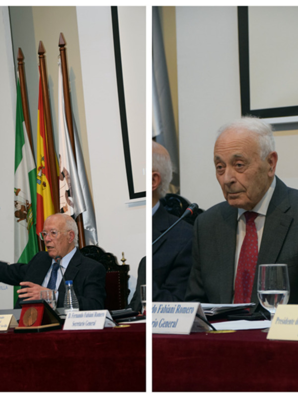 Dos hombres en trajes formales, uno hablando y el otro escuchando, en un entorno formal con banderas y un microfono en el frente.