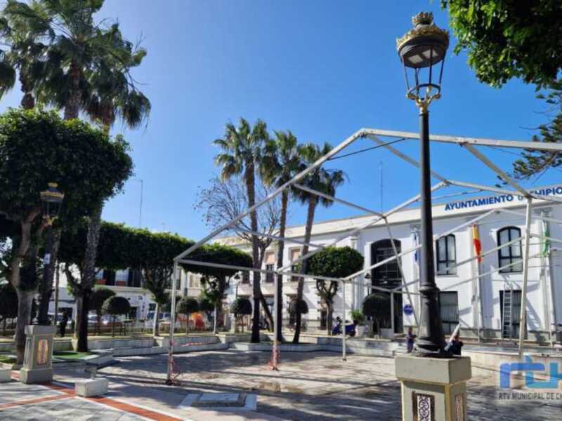 Plaza pública con palmeras y edificio municipal en el fondo, iluminada por una farola con una corona en el top.