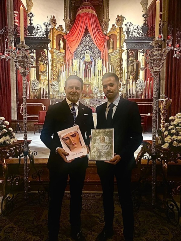 Dos hombres en trajes posan con un libro en un entorno decorado con velas y flores, posiblemente en una iglesia o iglesia.
