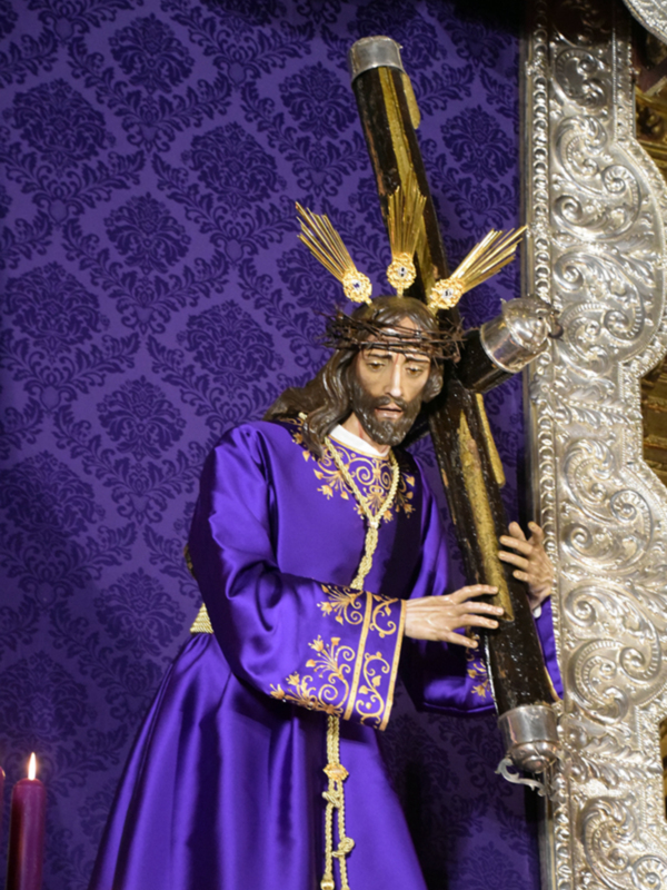 Estatua religiosa vestida de púrpura con un tocado dorado y un crucifijo grande, frente a un fondo de papel pintado en morado con detalles florales.