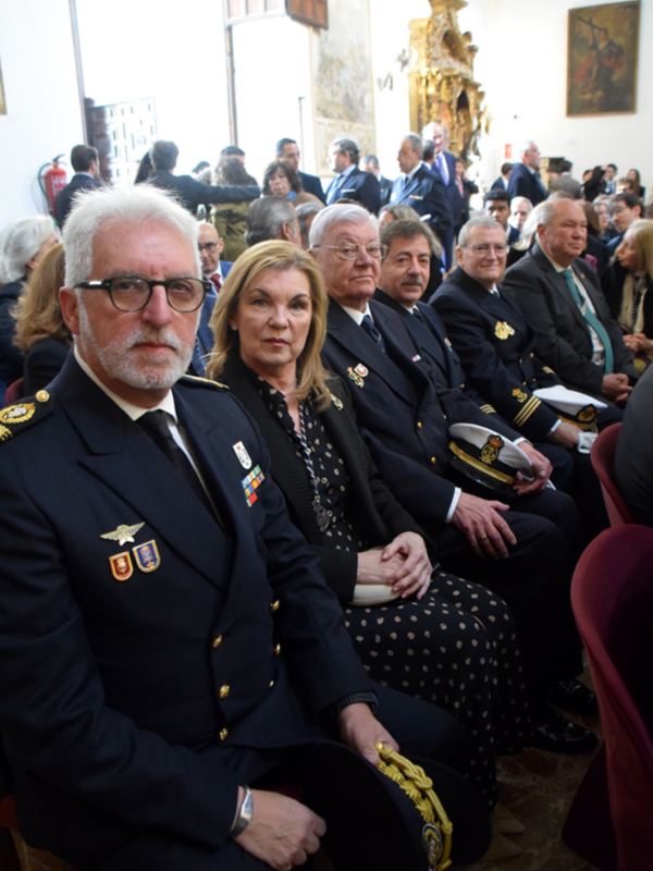 Asistentes en una ceremonia oficial, vestidos con uniformes y trajes formales, sentados en una fila en una sala decorada con arte y artefactos religiosos.