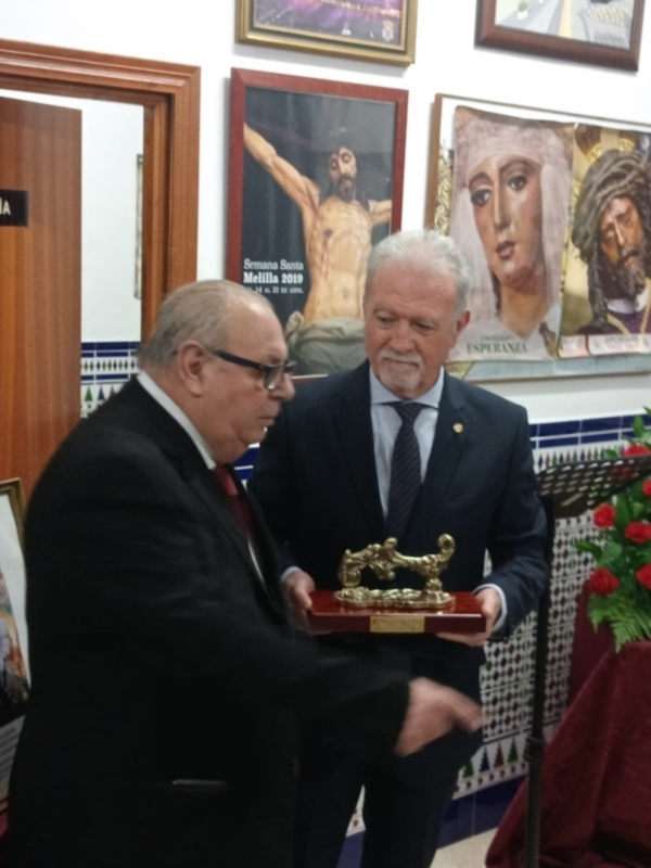 Dos hombres en traje intercambiando una placa de bronce en un ambiente formal con carteles religiosos en el fondo.