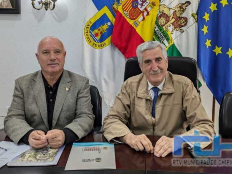 Dos hombres sentados en una mesa frente a banderas españolas y europeas, con un documento sobre la mesa.