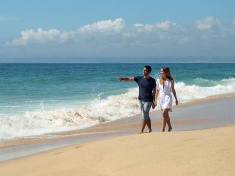 Un par disfruta de una caminata relajada por la playa, con olas rompiendo en la orilla y un cielo despejado de fondo.