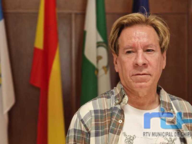 Un hombre con cabello dorado y barba, vistiendo una camisa a cuadros, frente a banderas de diferentes países.