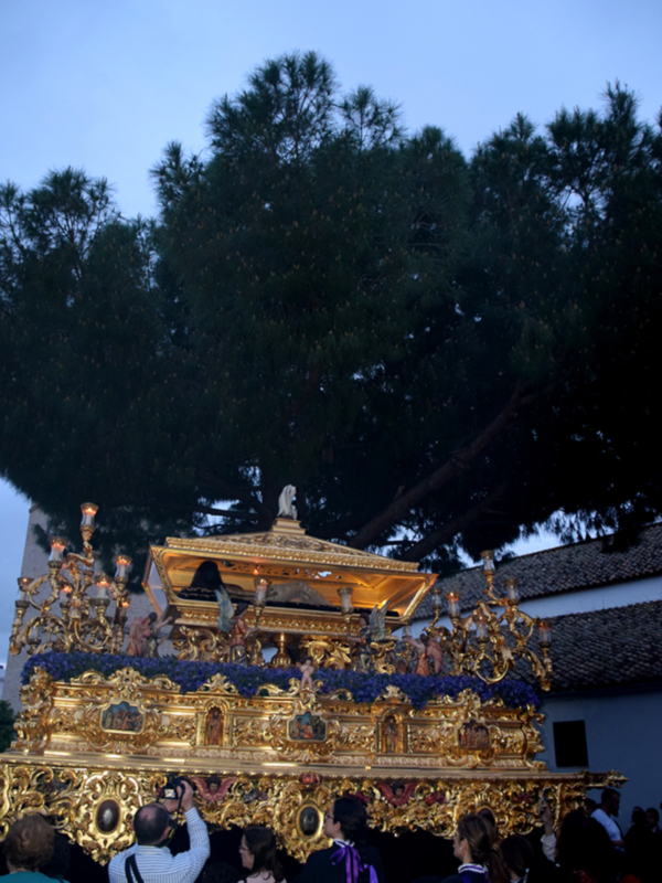 Procesión con paso ornado en oro y flores, bajo un árbol en la noche.