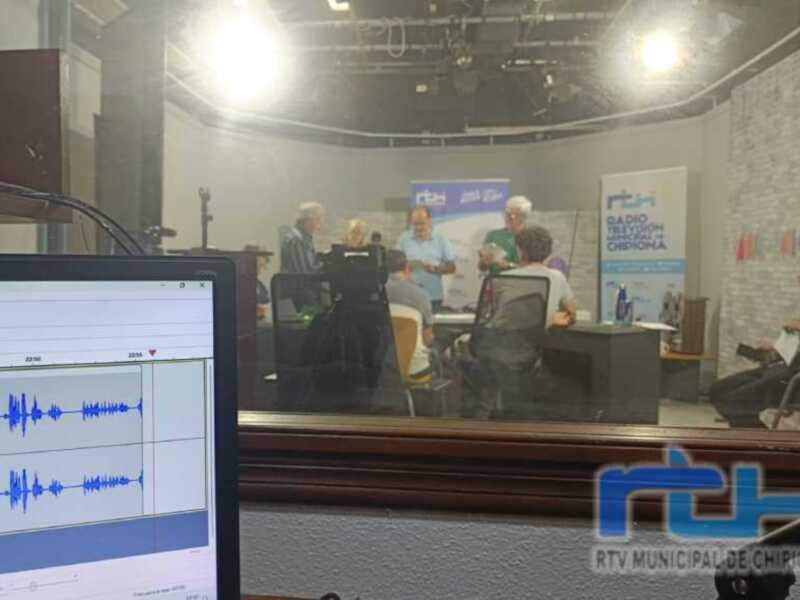 Un estudio de radio con personas hablando y un monitor en primer plano mostrando una señal de audio.