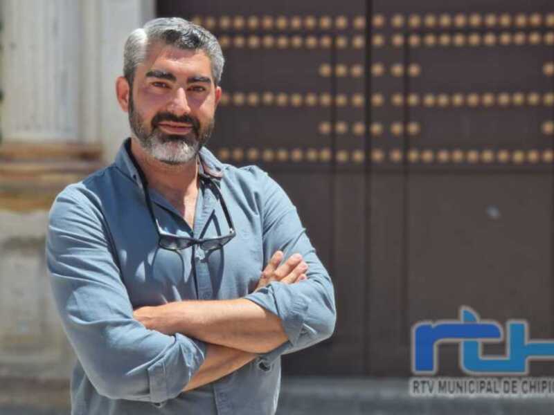 Un hombre con barba y gafas de sol, posando con los brazos cruzados en una calle con edificios de fondo.