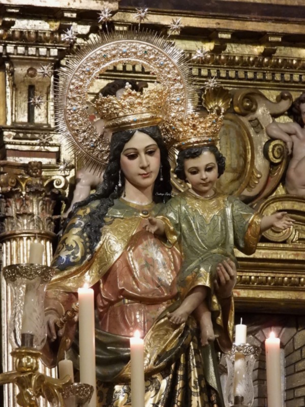 Escultura de la Virgen María con un niño en sus brazos, rodeada por detalles dorados y luces de velas.