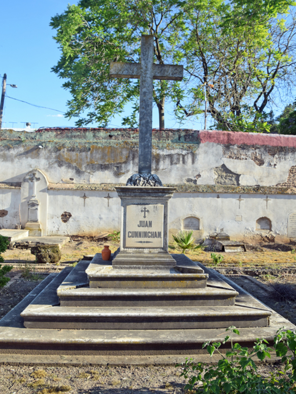 Un monumento sepulcral con la inscripción "JUAN CARRICHAM" en un cementerio, rodeado de vegetación y con una cruz en el centro.