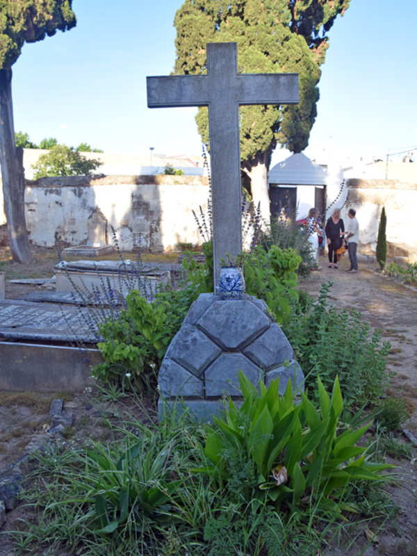 Un cementerio con una cruz de piedra en el centro, rodeada por vegetación y un hombre caminando cerca.