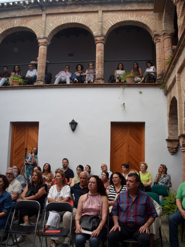 Una audiencia de personas sentadas en sillas, con un grupo de espectadores en la terraza superior observando el evento.