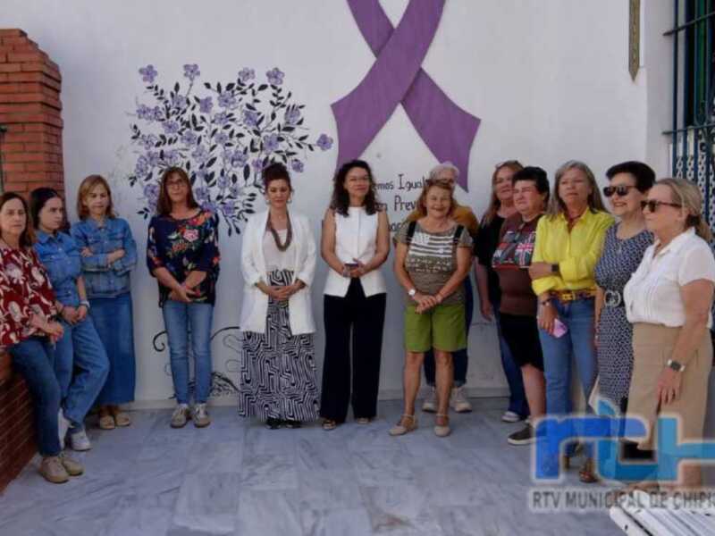 Una grupo de mujeres posando frente a una pared con un lazo morado, probablemente en apoyo al cáncer de mama.