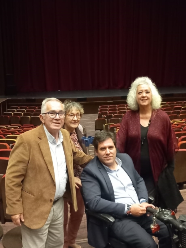 Un hombre en silla de ruedas se encuentra con dos personas en un teatro con cortinas rojas en el fondo.