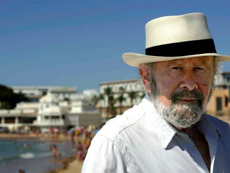 Un hombre con sombrero de paja y camisa blanca, posando frente a una playa con piscina y edificios en el fondo.