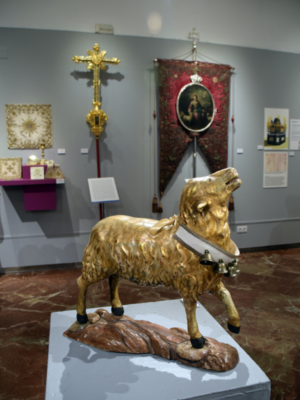 Un cuarto de exhibición con una estatua dorada de un cordero y un sepulcro en el suelo, junto a una cruz dorada y una bandera con un retrato.