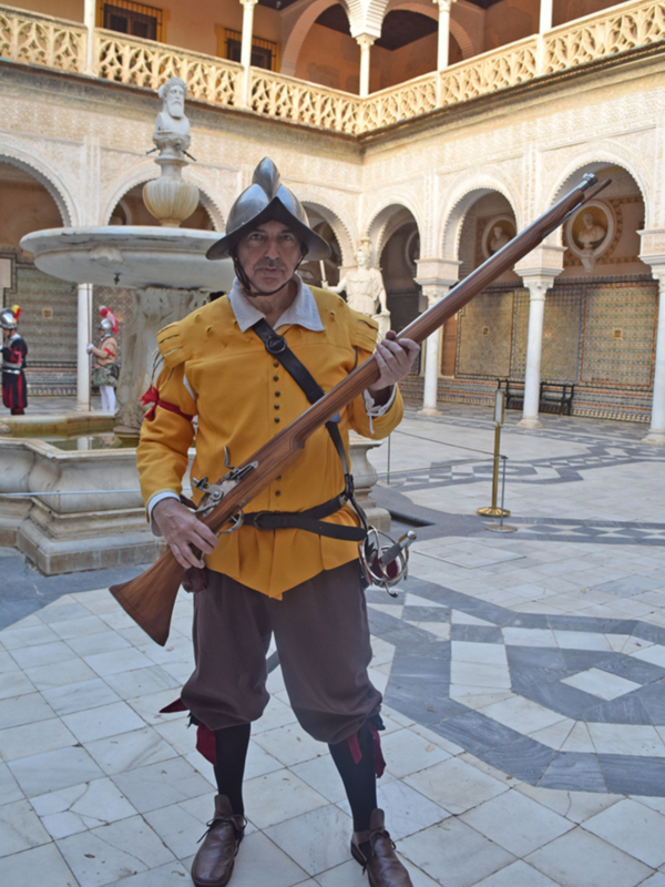 Un hombre vestido con traje histórico sostiene una armadura en un patio con arcos y columnas blancas.