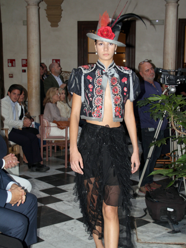 Desfile de moda con modelo en traje bordado y falda de encaje en un evento en un espacio con columnas clásicas.