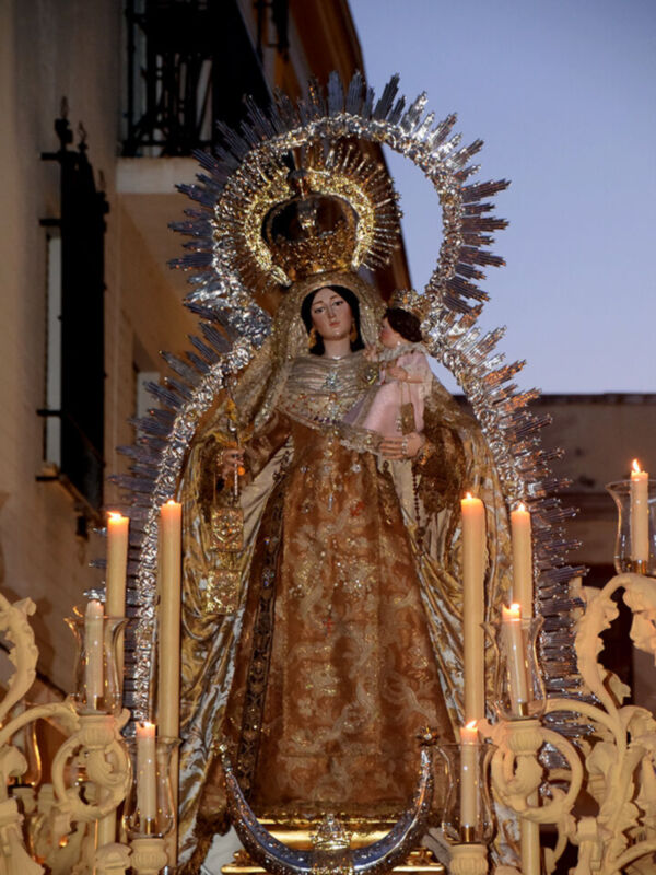 Procesión religiosa con imagen de la Virgen María adornada con velo y corona, rodeada de velas y ornamentos dorados.