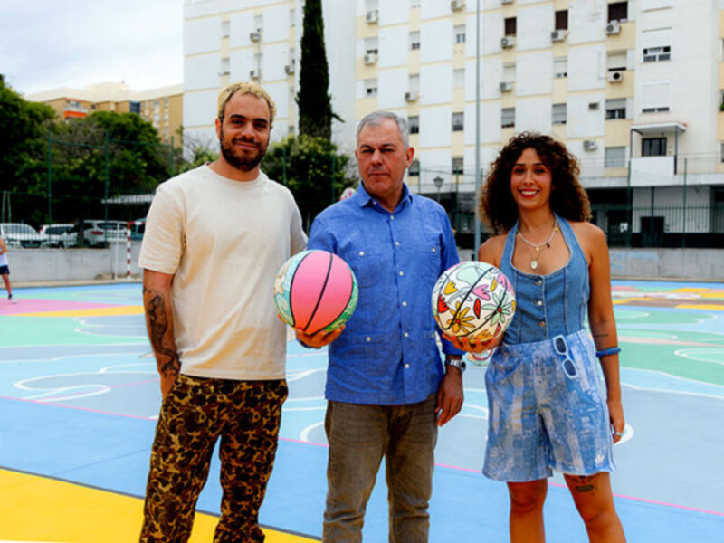 Tres personas posan con balones de baloncesto en una cancha de baloncesto al aire libre, con edificios residenciales en el fondo.