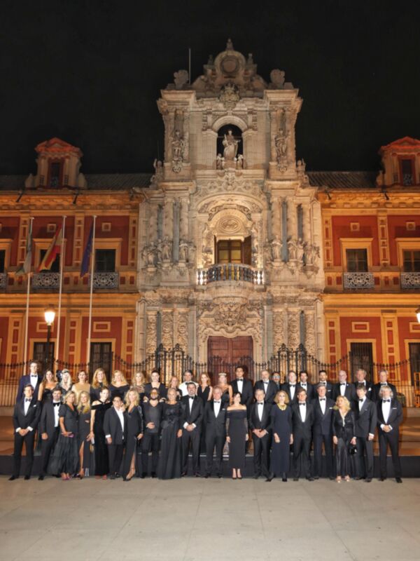 Una imagen de una gran fachada histórica con detalles arquitectónicos, donde un grupo de personas vestidas formalmente se encuentran en frente. La escena es nocturna y la iluminación refleja el ambiente serio y formal de los individuos.