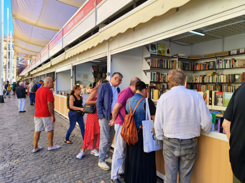 Una imagen de una librería al aire libre con gente comprando libros. La librería tiene estanterías llenas de libros y una tienda con toldos blancos. La gente está caminando por la calle mientras compran libros.