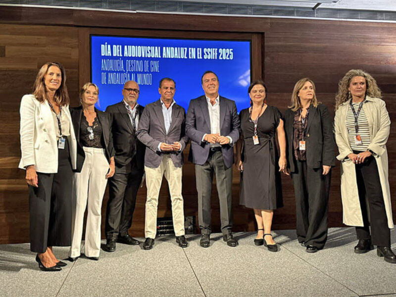 Se observa a una grupo de personas posando en un escenario con un fondo azul que dice "Día del Archivo Visual Andaluz en el SAIFF 2025". La persona de la izquierda lleva un traje blanco y pantalones negros, mientras que las demás están vestidas de manera formal.