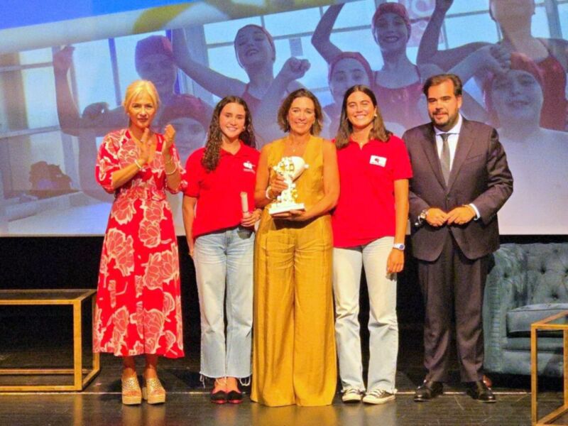 Una imagen de cuatro personas en un escenario, posiblemente en una ceremonia o evento. Dos mujeres jóvenes con camisetas rojas sostienen un trofeo mientras dos mujeres más viejas las miran. Un hombre en traje está a su derecha, y detrás de ellos hay una pantalla con imágenes de mujeres en trajes de baño.