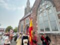 Desfile histórico frente a una iglesia con arcos y vitrales, personas vestidas en trajes antiguos sostienen banderas.